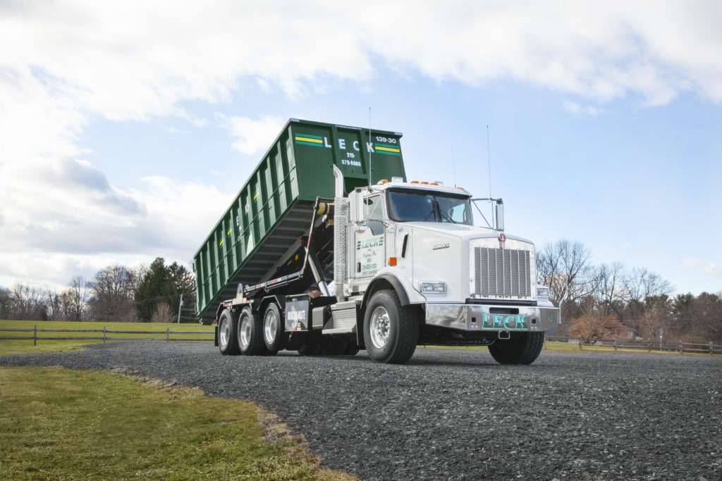 A Leck truck with roll-off dumpster on the back of it is pictured with a dumpster securely fastened onto its back, and the rails are raised high into the air, indicating readiness to unload the dumpster. The truck's hydraulic system is engaged, preparing to tilt the dumpster backward for unloading. This scene depicts a typical operation of a roll-off truck in the process of emptying a dumpster.