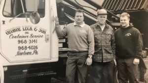 An antiqued-style photo depicts Walter and George Leck, the founders of Leck Waste, standing proudly in front of a vintage truck. Both men wear work attire indicative of the era, suggesting the photo's historical significance. Their expressions reflect determination and vision as they stand beside their truck, symbolizing the beginnings of their waste management enterprise.