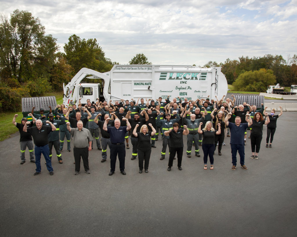 Leck Waste in action company group shot, approximately fifty employees stand in front of a front-load garbage truck, arms raised in celebration. The truck is flanked by two front-load containers on each side. The group is likely celebrating a successful waste management operation or milestone achievement in the sanitation industry.