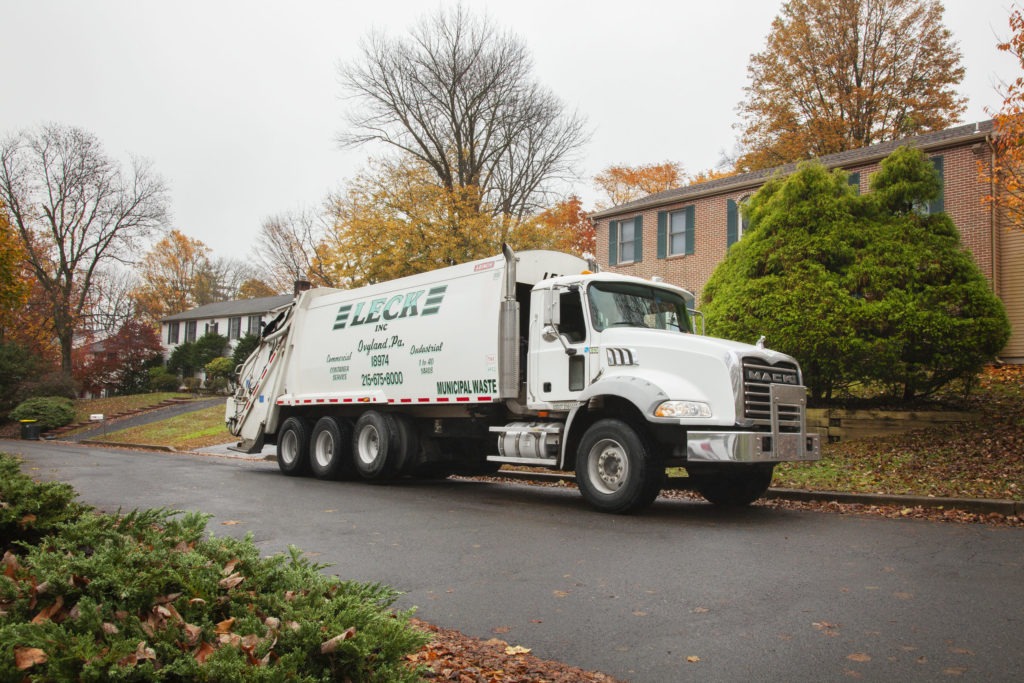 A Leck rear-load garbage truck, a Mack Granite model with a Leach body, is parked in a residential neighborhood on a crisp fall day. Colorful houses line the street in the background, surrounded by trees displaying autumn foliage. The truck's presence signifies the routine residential waste collection service provided by Leck Waste, blending into the familiar scene of suburban life.