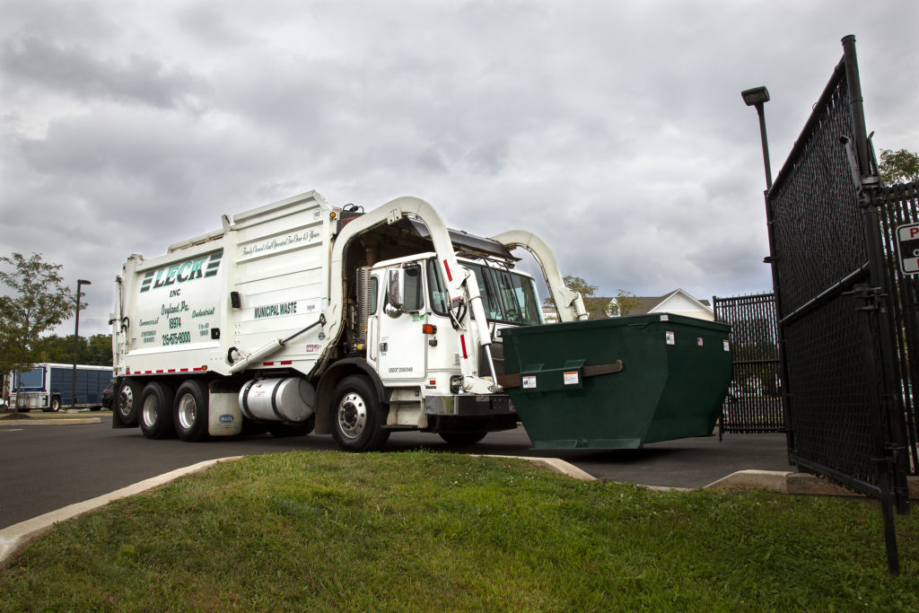 Leck trash truck picking up dumpster, A front-load garbage truck maneuvers within a gated enclosure, lifting a large container into its compactor. The truck's hydraulic arms are extended, securely gripping the container as it prepares to lift it for disposal. The scene illustrates a typical waste collection process within a controlled access area.