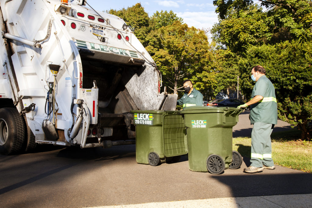 Two Leck Waste Services employees in safety clothing stand beside a rear-load garbage truck, each holding a trash cart. One cart is already tilted into the truck's compactor, while the other is being maneuvered towards the opening. The truck's rear door is open, ready to receive the carts. This scene captures the routine task of waste collection by municipal workers, ensuring proper disposal of refuse into the garbage truck.